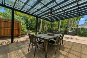 Eine überdachte Terrasse mit Tisch und Stühlen, umgeben von üppiger Bepflanzung und einem Holzzaun.