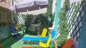 Une terrasse avec une pelouse artificielle verte, une table recouverte d’une nappe à motifs floraux et des fauteuils de jardin aux couleurs vives.