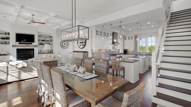 A spacious living room with a fireplace, a dining area with wicker chairs, and a kitchen with white cabinets and a large island. - Stunning Luxury Beach Home: Gulf Views, Large Private Pool, Free Beach Setup! (Panama City Beach)