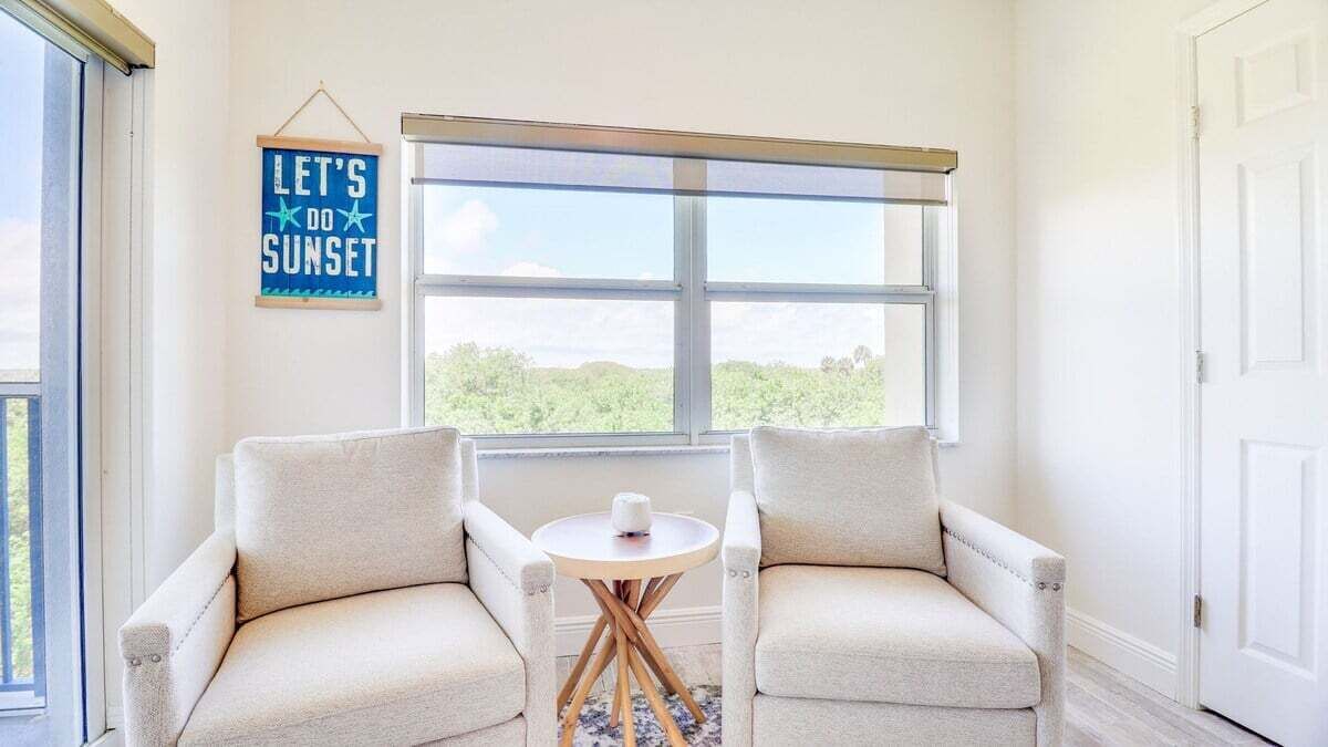 Una habitación con dos sillones color beige orientados hacia una ventana que ofrece vistas de zonas verdes. Una pequeña mesa redonda con un objeto blanco en el centro. Un cartel en la pared dice 'LET'S DO SUNSET'.