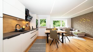 A modern kitchen with white cabinets, a black countertop, and a wooden backsplash. There is a dining area with a table and chairs, and a living area with a sofa and a wall-mounted artwork. - Ferienwohnung Silvia , Mondsee Zentrum (Mondsee)