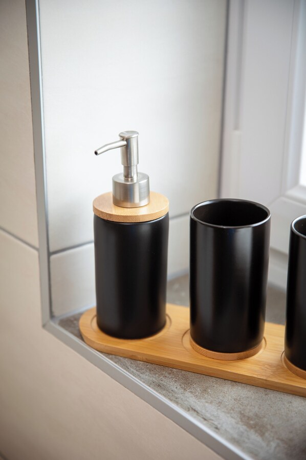 Un baño con espejo, un dispensador de jabón negro sobre una bandeja de madera y dos tazas negras en el mostrador.