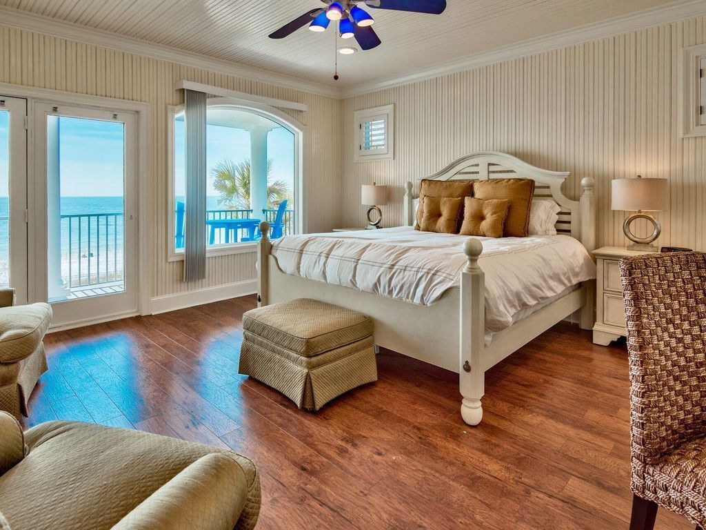 A bedroom with a large bed, a ceiling fan, a view of the beach, and a balcony.
