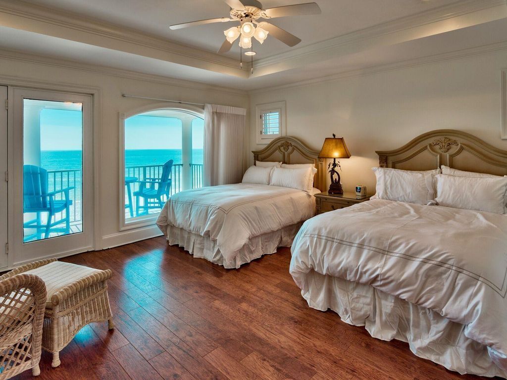 Ein Hotelzimmer mit zwei Betten, einem Deckenventilator, einem Nachttisch mit Lampe, einem Korbstuhl und Blick auf das Meer durch große Fenster.