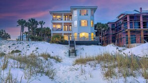 A multi-story beachfront property with a staircase leading to the entrance, surrounded by sand and sparse vegetation. - Stunning Views! 9BR Gulf Front Home, Pool, Elevator, Pool Table (Destin)