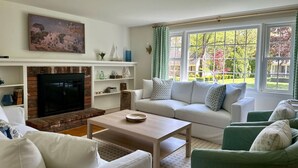Un salon avec une cheminée, des canapés blancs, une table basse et une grande fenêtre avec des rideaux.