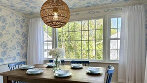 Une salle à manger avec une table en bois dressée pour six personnes, des chaises bleues, un luminaire suspendu et une fenêtre ornée de rideaux blancs.