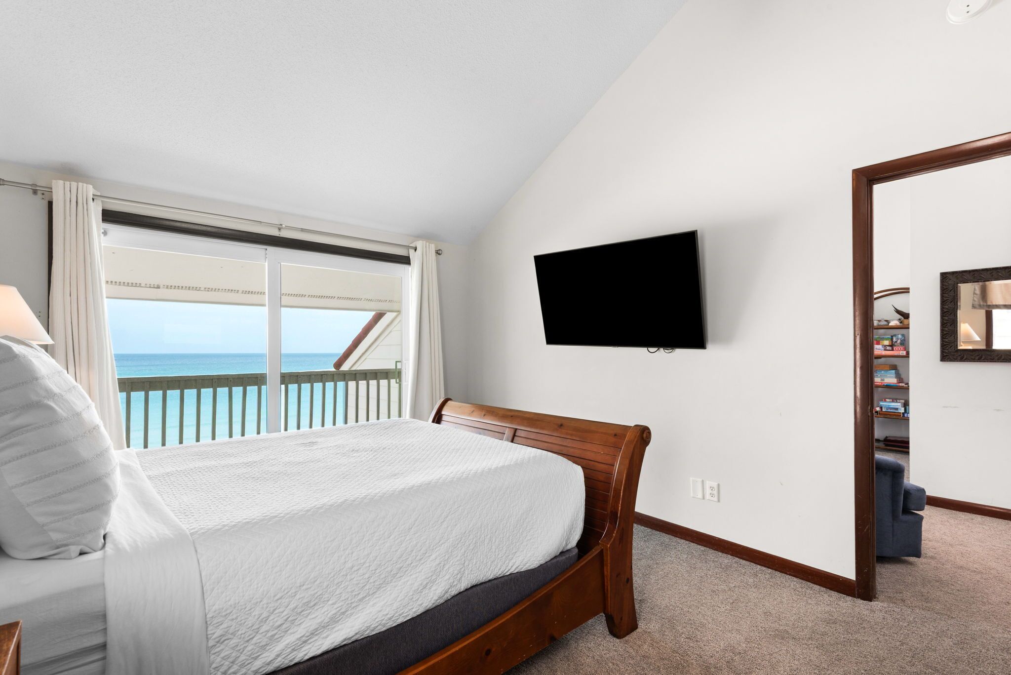 A bedroom with a bed, a TV mounted on the wall, a window with curtains, and a view of the ocean.