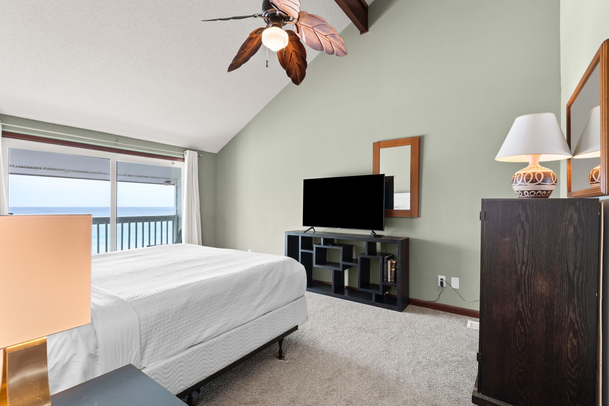 A bedroom with a bed, a TV, a ceiling fan, a dresser, and a window with a view.