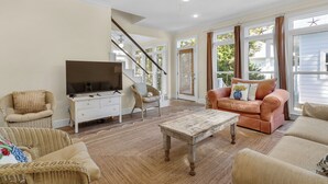Ein geräumiges Wohnzimmer mit Fernseher, bequemen Sitzgelegenheiten, einer Treppe und großen Fenstern.