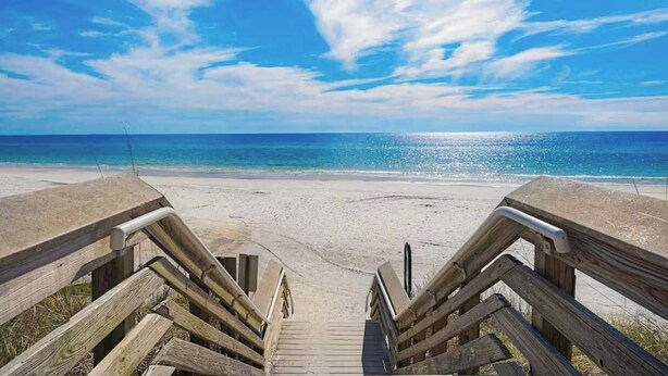Ein hölzerner Steg führt zu einem Strand mit blauem Himmel und Meer.