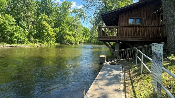 A wooden cabin with a deck overlooking a river, surrounded by lush greenery. - 6 Bed River Home, Right On St. Joseph River. (Union City)