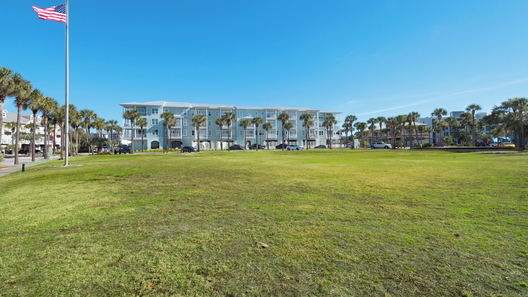 Un edificio grande de varios pisos con techo plano y balcones, rodeado de césped y palmeras.
