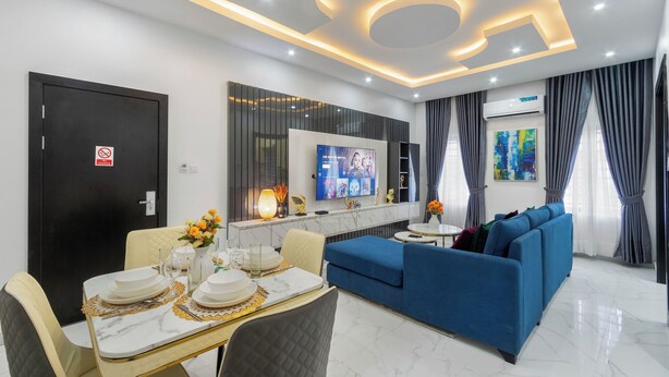 A modern living room with a blue sofa, a dining table set with plates and a vase of flowers, and a TV mounted on the wall.