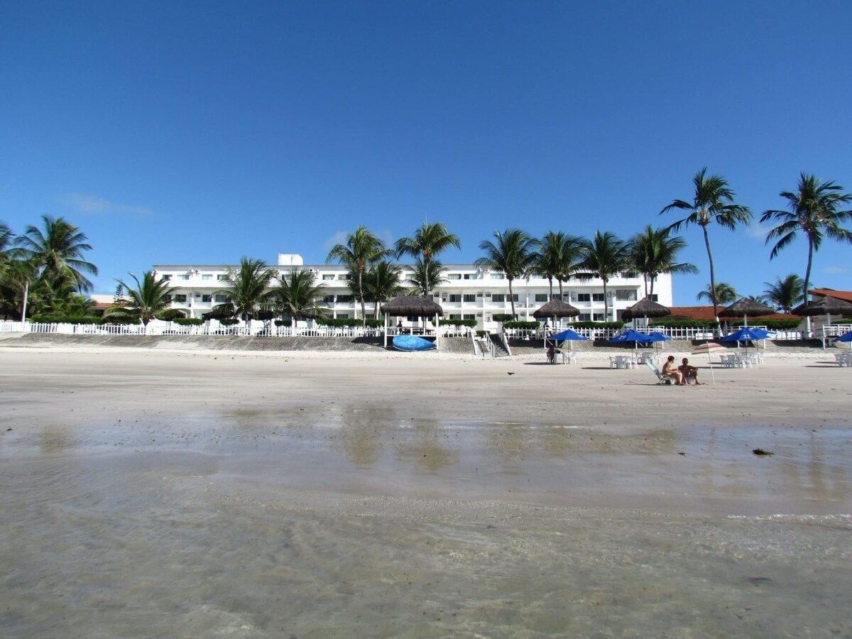 A beach with a hotel in the background, palm trees, and umbrellas.