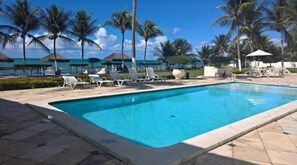 Una piscina con sillas de descanso, palmeras y un cielo azul despejado.