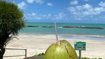 A green coconut with a straw on a wooden table at a beach with a clear sky and ocean in the background.
