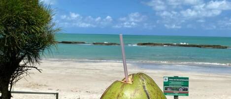 A green coconut with a straw on a wooden table at a beach with a clear sky and ocean in the background.