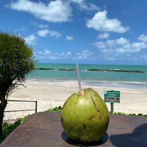 Un coco verde con una pajita sobre una mesa de madera en una playa, con cielo despejado y océano al fondo.