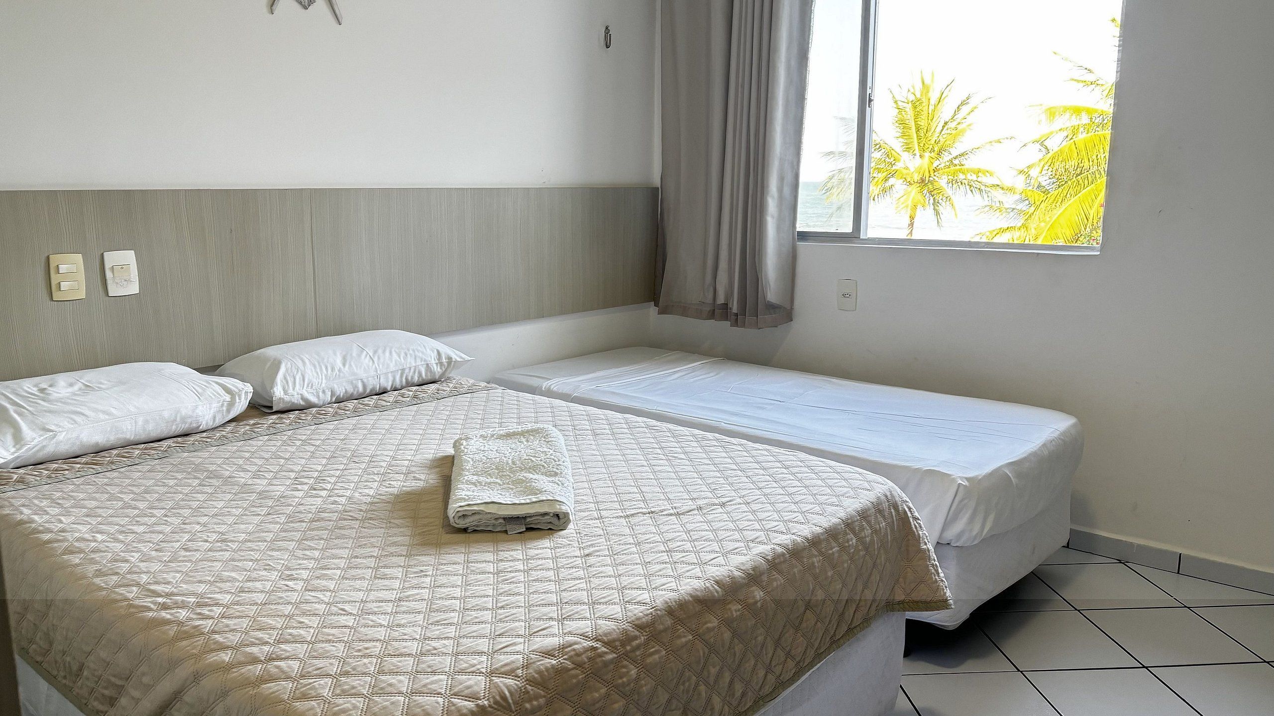 A hotel room with a large bed, a smaller bed, a window with a view of palm trees, and a curtain.