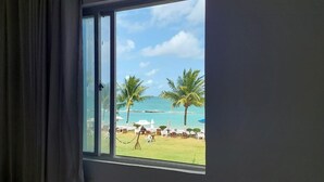 A room with a large window offering a view of a beach with palm trees and a clear sky. - Marinas Tamandaré próximo à Carneiros  (Tamandaré)