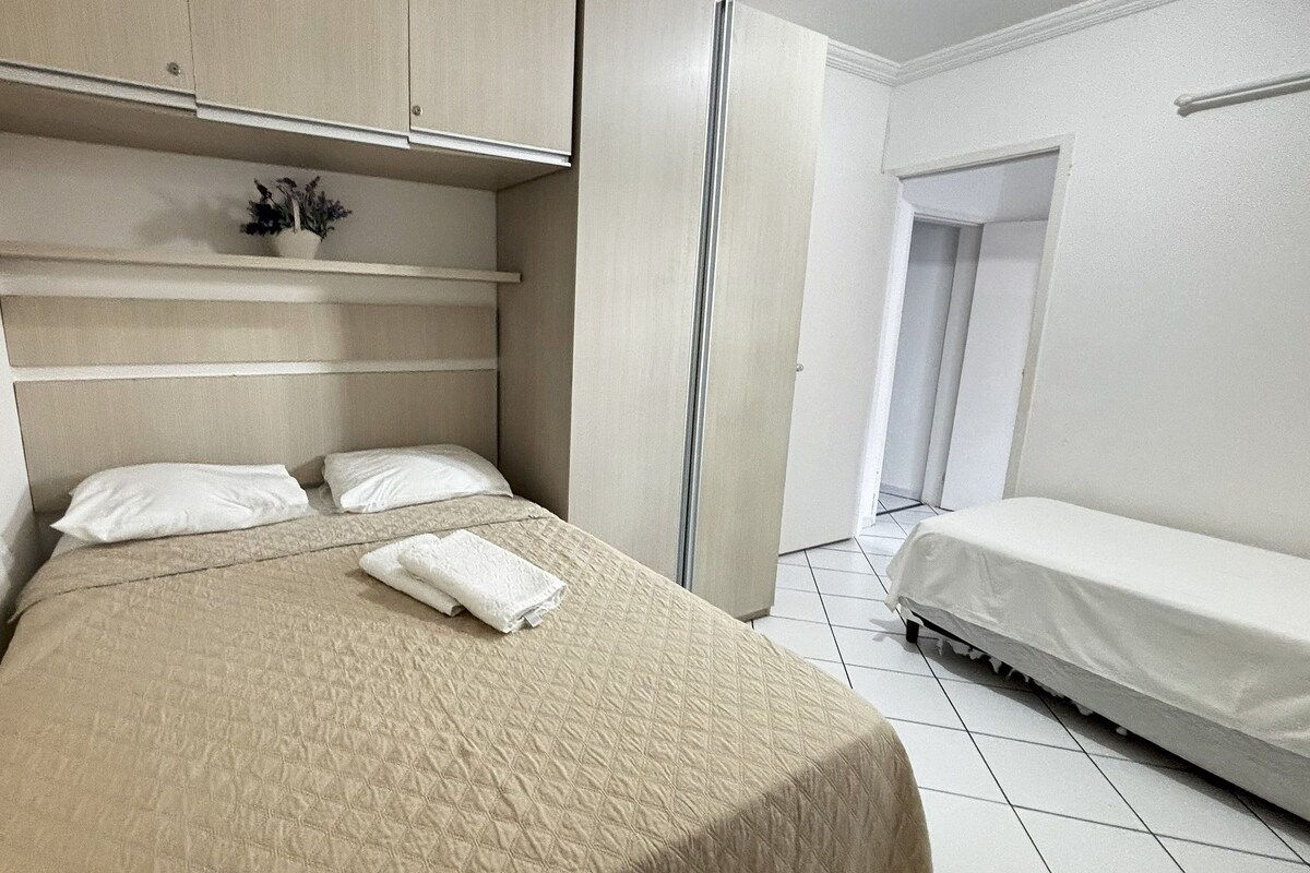 A bedroom with a bed, a bedside table, a small potted plant, and a door leading to another room.