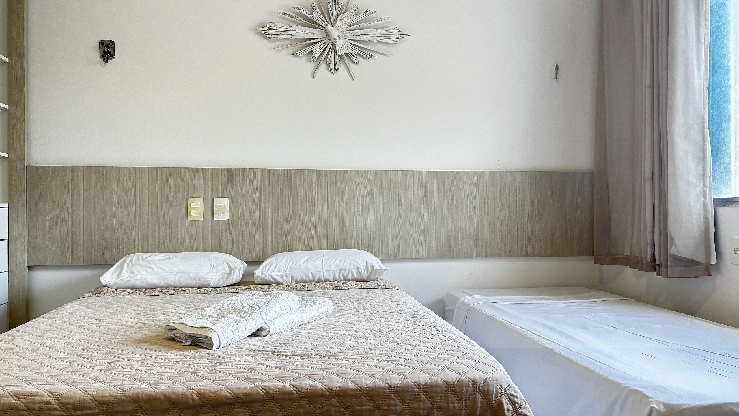 Habitación de hotel con cama, mesita de noche, lámpara de pared, un adorno en la pared y una ventana con cortinas.