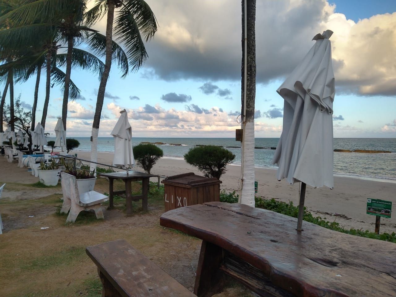 A beachside seating area with white umbrellas, wooden tables, and chairs, overlooking the ocean.