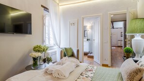 Une chambre à coucher comprenant un lit, une télévision, une fenêtre avec des rideaux, une table de chevet avec des serviettes et une vue sur une autre pièce.