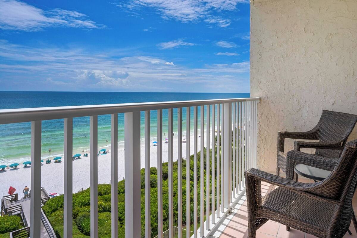 A balcony with wicker chairs overlooking a beach with umbrellas and people.