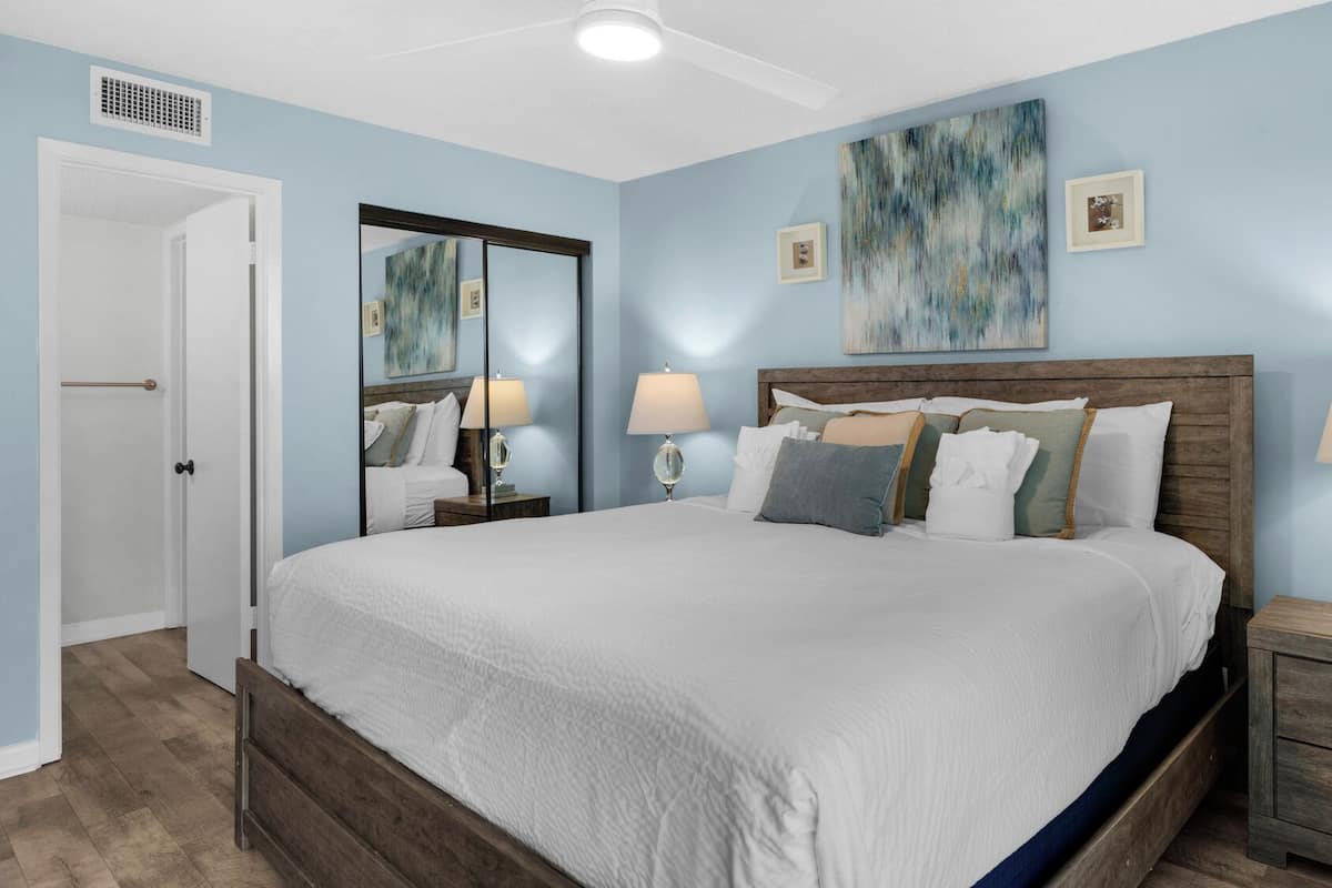 A bedroom with a large bed, two bedside tables with lamps, a wooden headboard, a large mirror, and a ceiling fan.