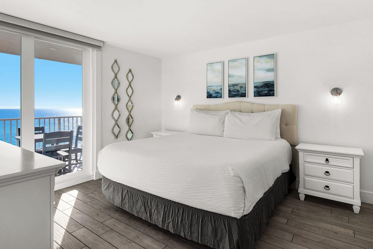 A bedroom with a large bed, a nightstand, a dresser, and a view of the ocean.