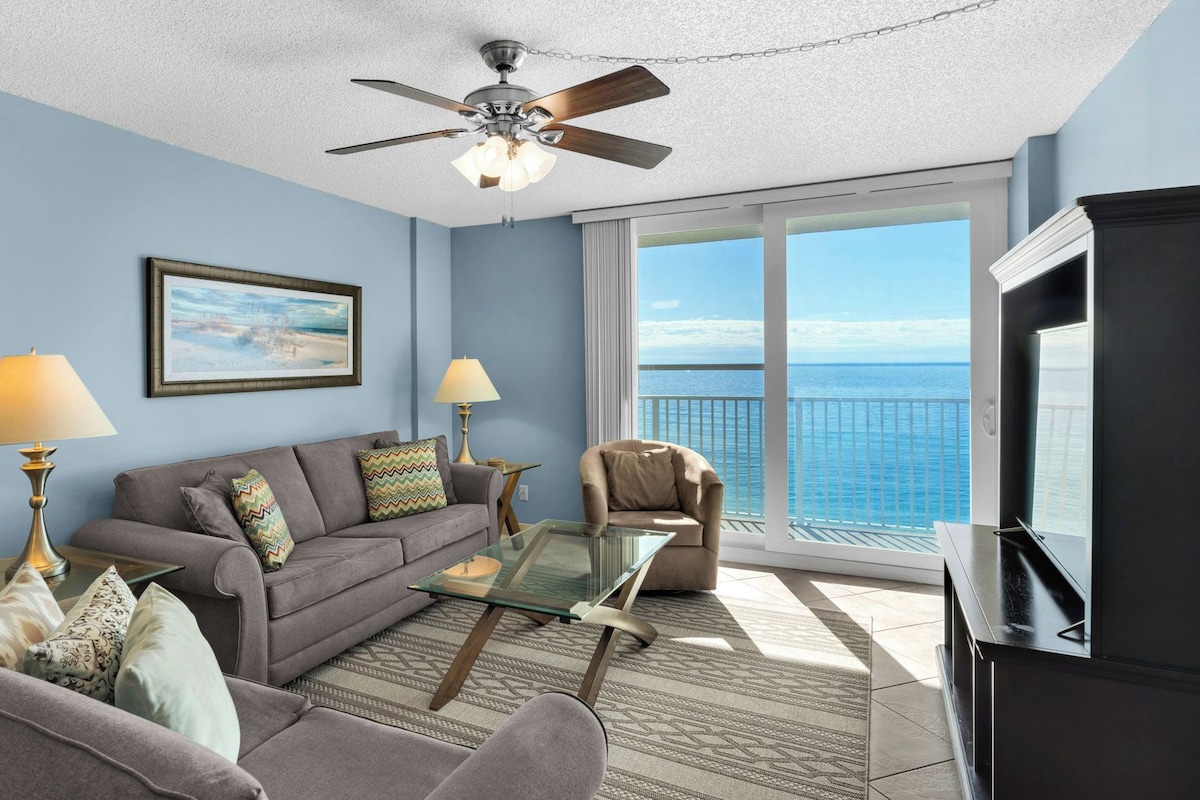 A living room with a sofa, two lamps, a glass coffee table, a TV, and a balcony with a view of the ocean.