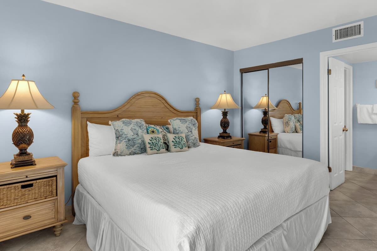 A bedroom with a large bed, two bedside tables with lamps, a wooden nightstand, and a mirror on the wall.