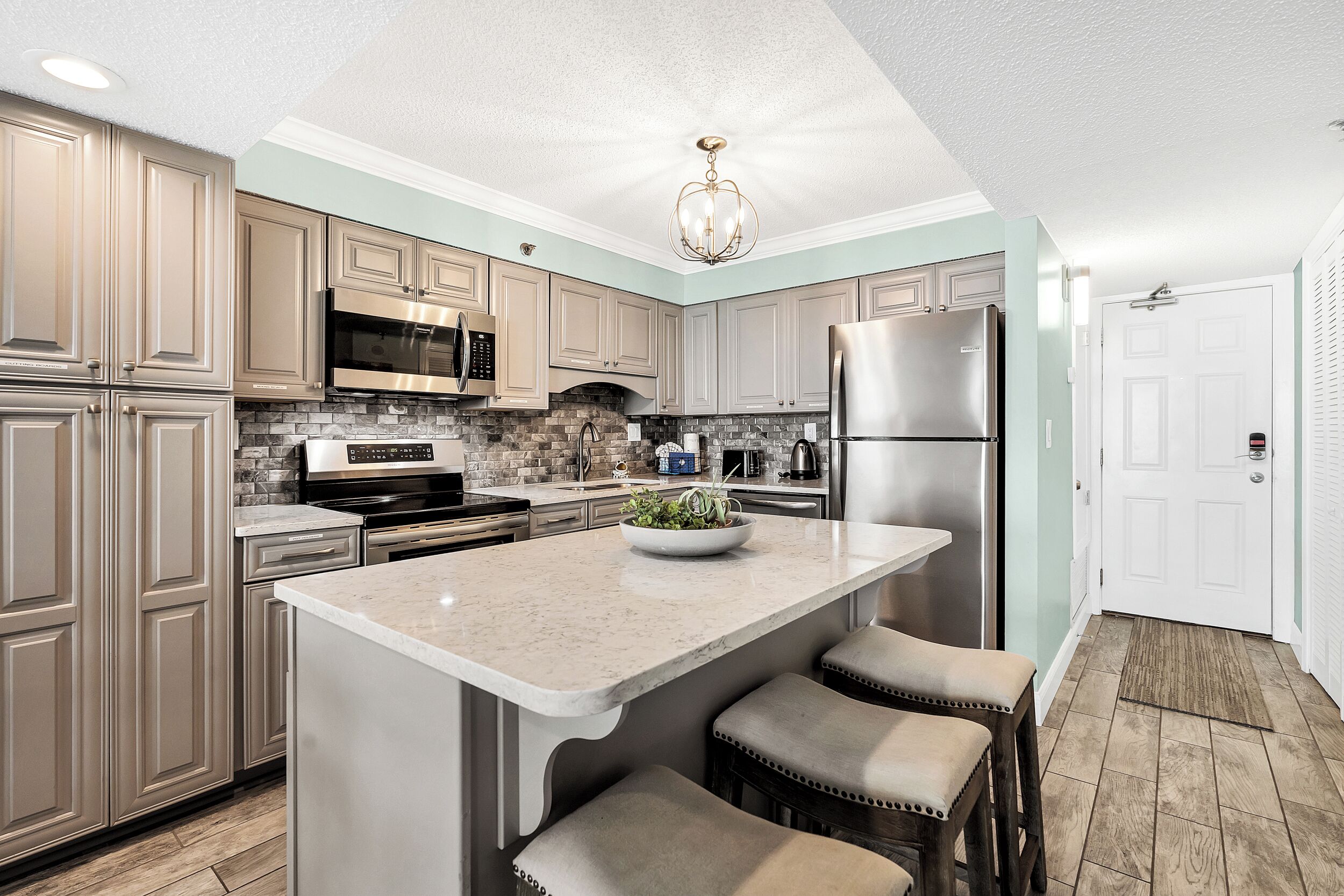 Une cuisine moderne avec des meubles de couleur beige, des appareils en acier inoxydable, un îlot central doté de tabourets et un mur d’accent d’une teinte turquoise claire.
