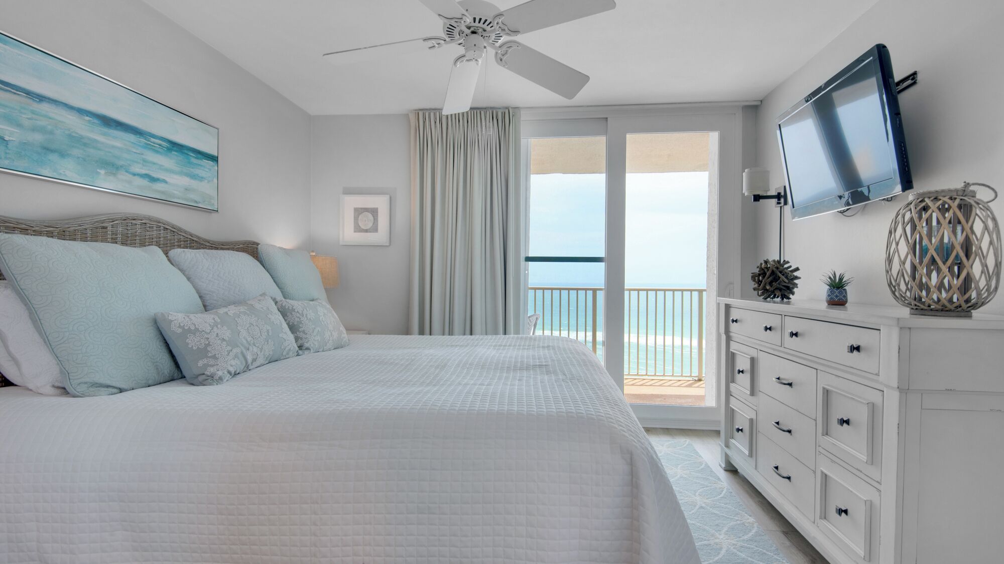 A bedroom with a bed, a ceiling fan, a TV, and a view of the ocean.