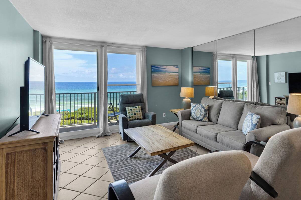 A living room with a view of the ocean, a flat-screen TV, a sofa, armchairs, and a coffee table.