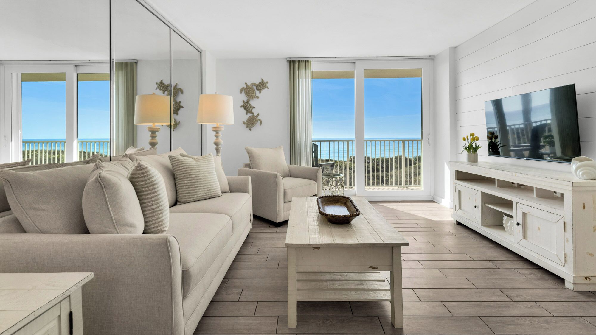 A modern living room with a sofa, coffee table, and TV. There are large windows with a view of the sea.