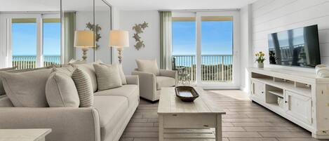 A modern living room with a sofa, coffee table, and TV. There are large windows with a view of the sea.