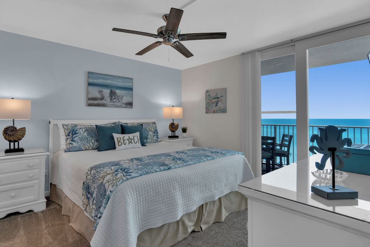 A bedroom with a bed, two bedside tables, a ceiling fan, and a view of the ocean through a large window.
