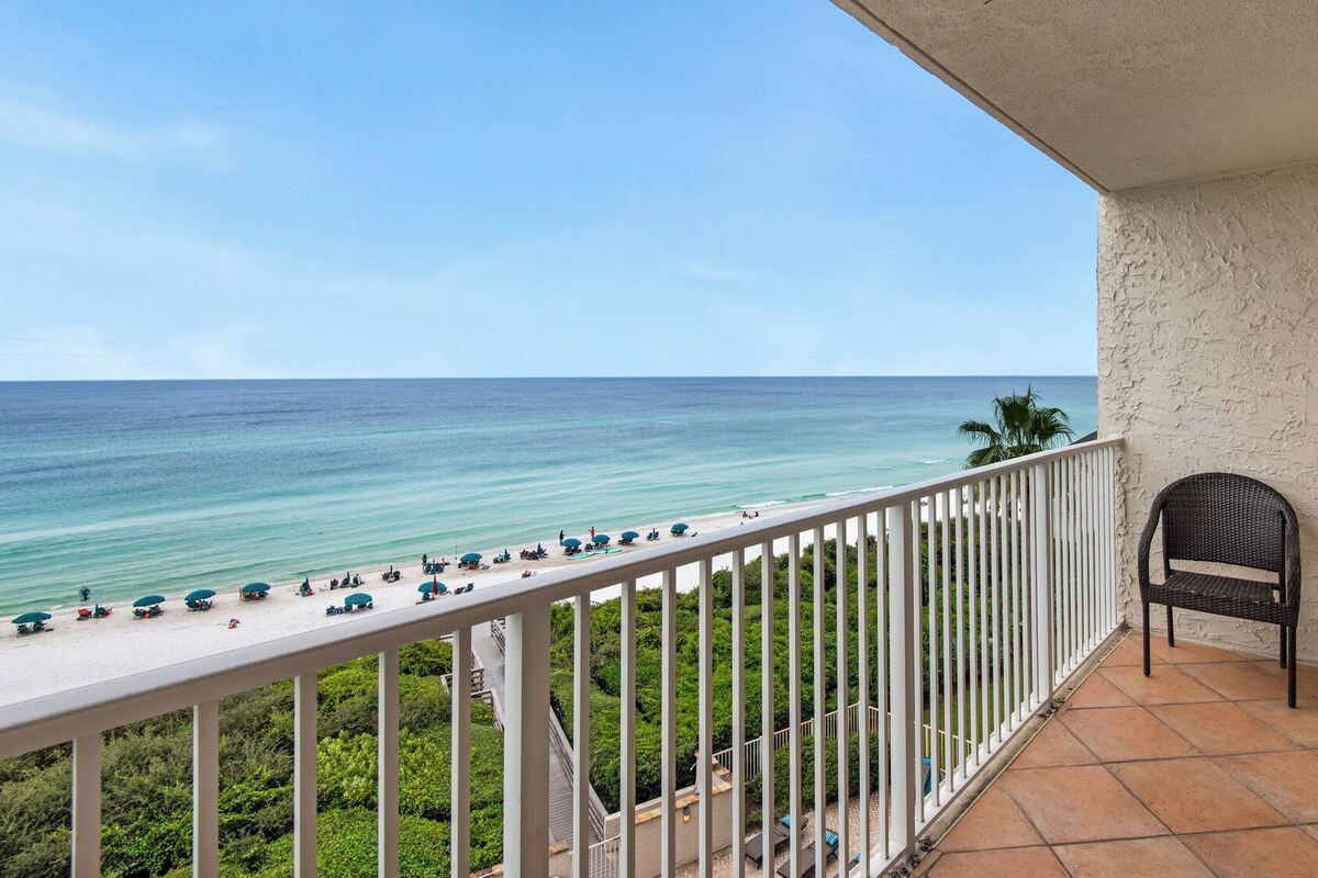 A balcony with a view of the beach and ocean. There are several umbrellas and lounge chairs on the beach.