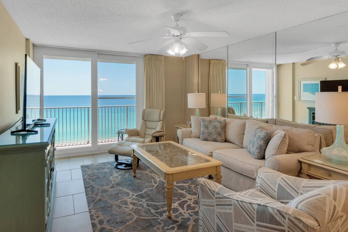 A spacious living room with a large window offering a view of the ocean, a ceiling fan, and a sofa with patterned cushions.