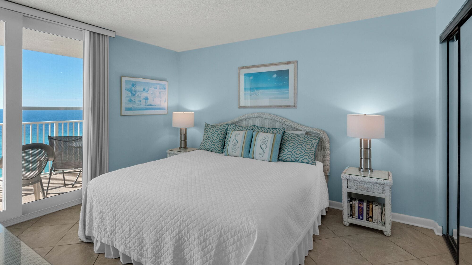 A bedroom with a large bed, two bedside tables with lamps, a small bookshelf, and a balcony with a chair.