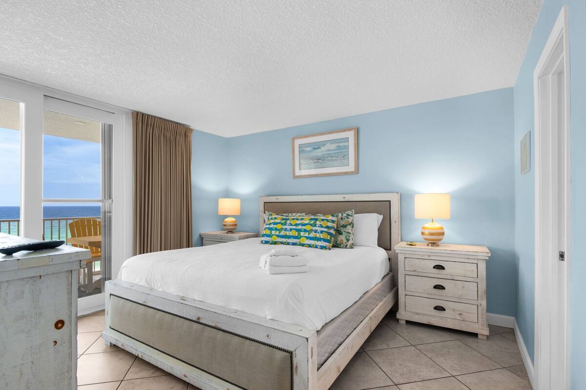 A bedroom with a bed, two bedside tables, a nightstand, a chair, and a view of the ocean.