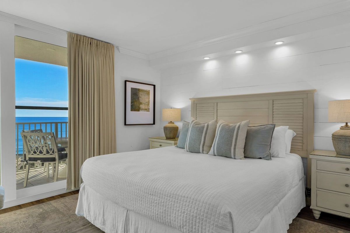 Un dormitorio amplio con una cama grande, una mesita de noche con lámpara, una silla y vista al océano.