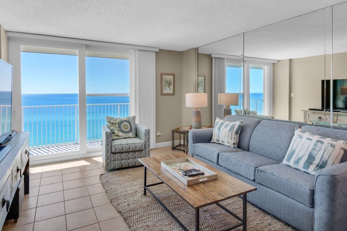 A living room with a large TV, a sofa, a chair, and a coffee table. There is a balcony with a view of the ocean.