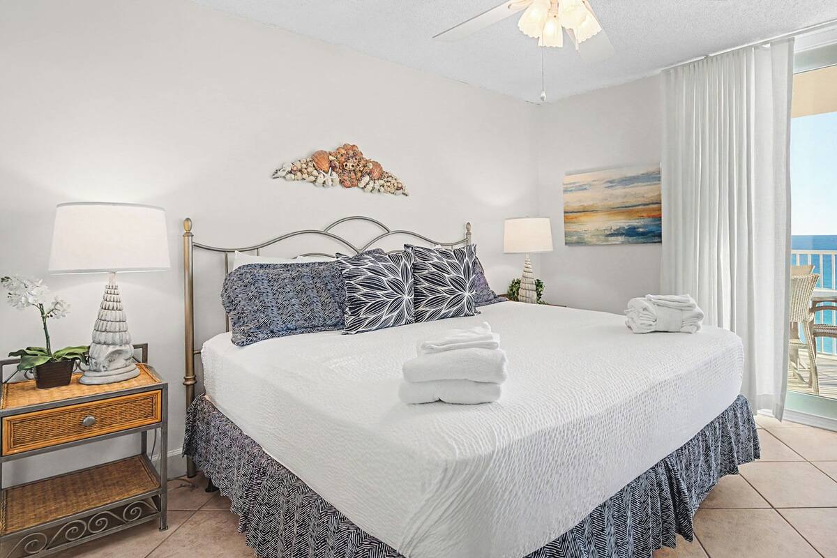 A hotel room with a bed, two bedside tables, a nightstand, a potted plant, a painting, and a balcony with a view of the sea.