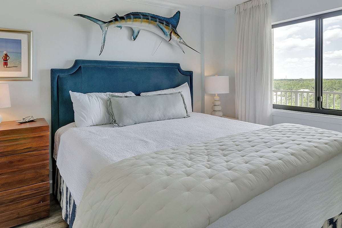 A hotel room with a large bed, a blue headboard, a framed picture on the wall, and a view of the outdoors through the window.