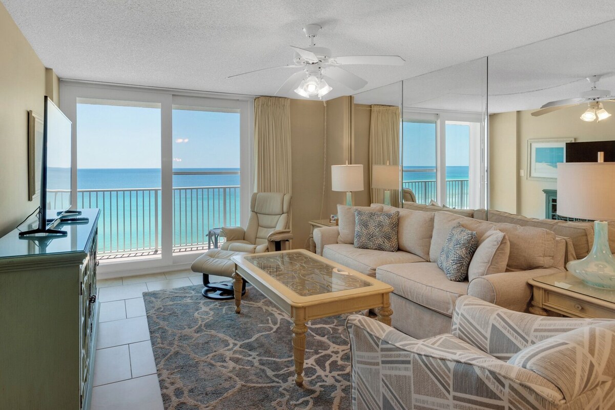 A spacious living room with a large window offering a view of the ocean, a ceiling fan, and a sofa with patterned cushions.