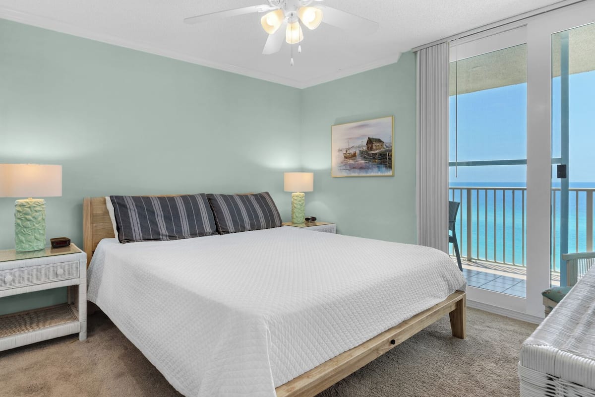 A bedroom with a bed, two bedside tables with lamps, a painting, and a view of the ocean.